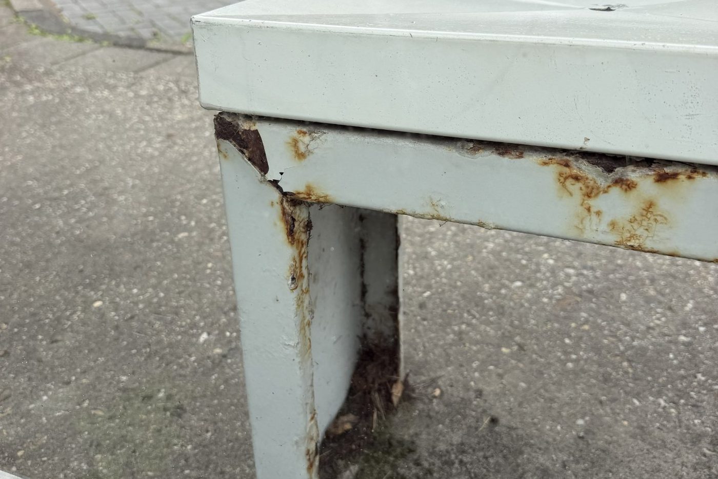 Hoek van een stalen bankje in een bushokje, met roest en afbladderende coating vanuit de randen
