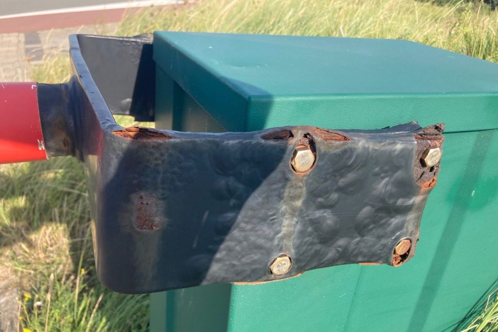 Zwarte beugel van een slagboom met doorgeroeste randen rond de RVS bouten, op een groene behuizing